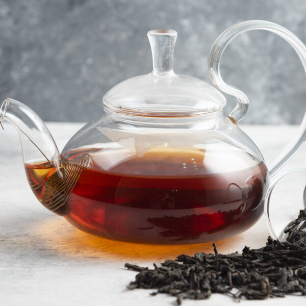 Teekanne mit aufgebrühtem Tee. Neben der Kanne liegen getrocknete Teepflanzen, die sich aus einem liegenden Behälter auf einen Tisch verteilen.